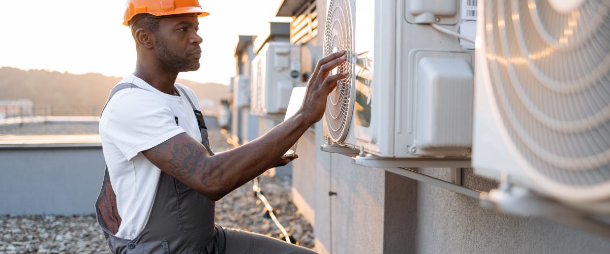 Side view of skilled man typing on wireless laptop while kneeling near hanging air conditioner outdoors. African american specialist using modern technologies for quickly repairing of cooling system.