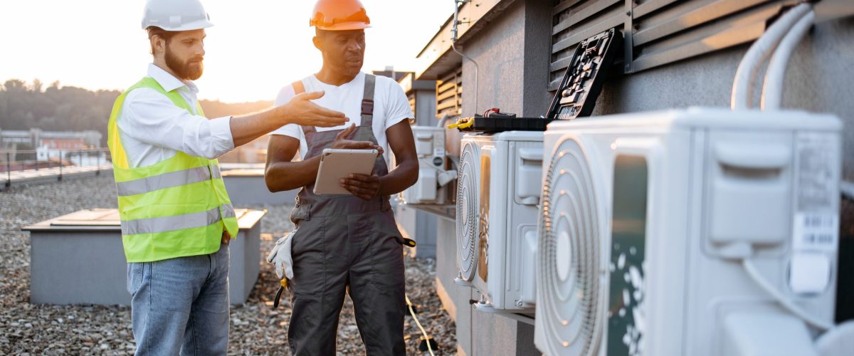 Multicultural males advising with each other about repairing of air conditioner while standing near broken cooling system outdoors. Men using tablet for scrolling websites with useful information.